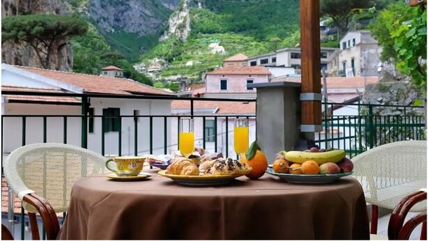 Una mesa servida con alimentos para el desayuno, como cruasanes, fruta y jugo, con vistas a un pueblo montañoso de arquitectura tradicional.