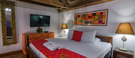 Quarto de hotel com teto de madeira, uma cama grande com colcha vermelha e branca, uma televisão na parede, um espelho decorativo e um colcha colorida pendurada sobre a cama.