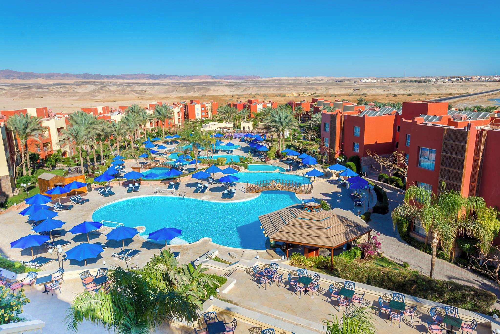 Un resort con una piscina grande rodeada de tumbonas y sombrillas, palmeras y un paisaje desértico al fondo.