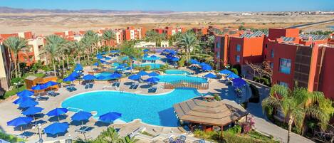 Un resort con una piscina grande rodeada de tumbonas y sombrillas, palmeras y un paisaje desértico al fondo.