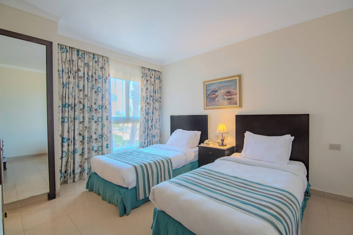 A hotel room with two beds, a nightstand with a lamp, a painting on the wall, and a window with curtains.
