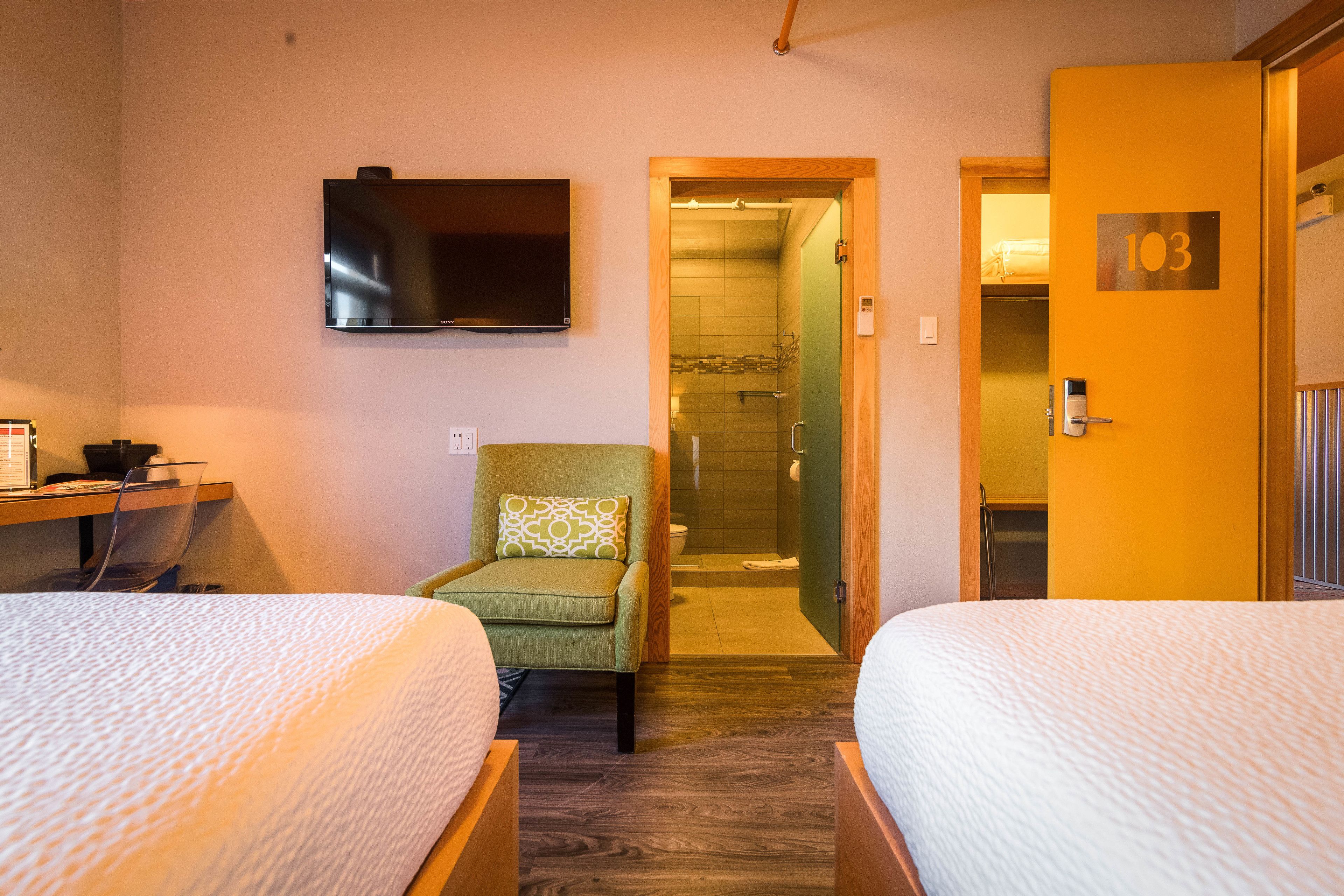 Ein Hotelzimmer mit zwei Betten, einem Fernseher, einem Sessel mit Kissen, einer Duschkabine und einer Tür mit der Nummer 103.