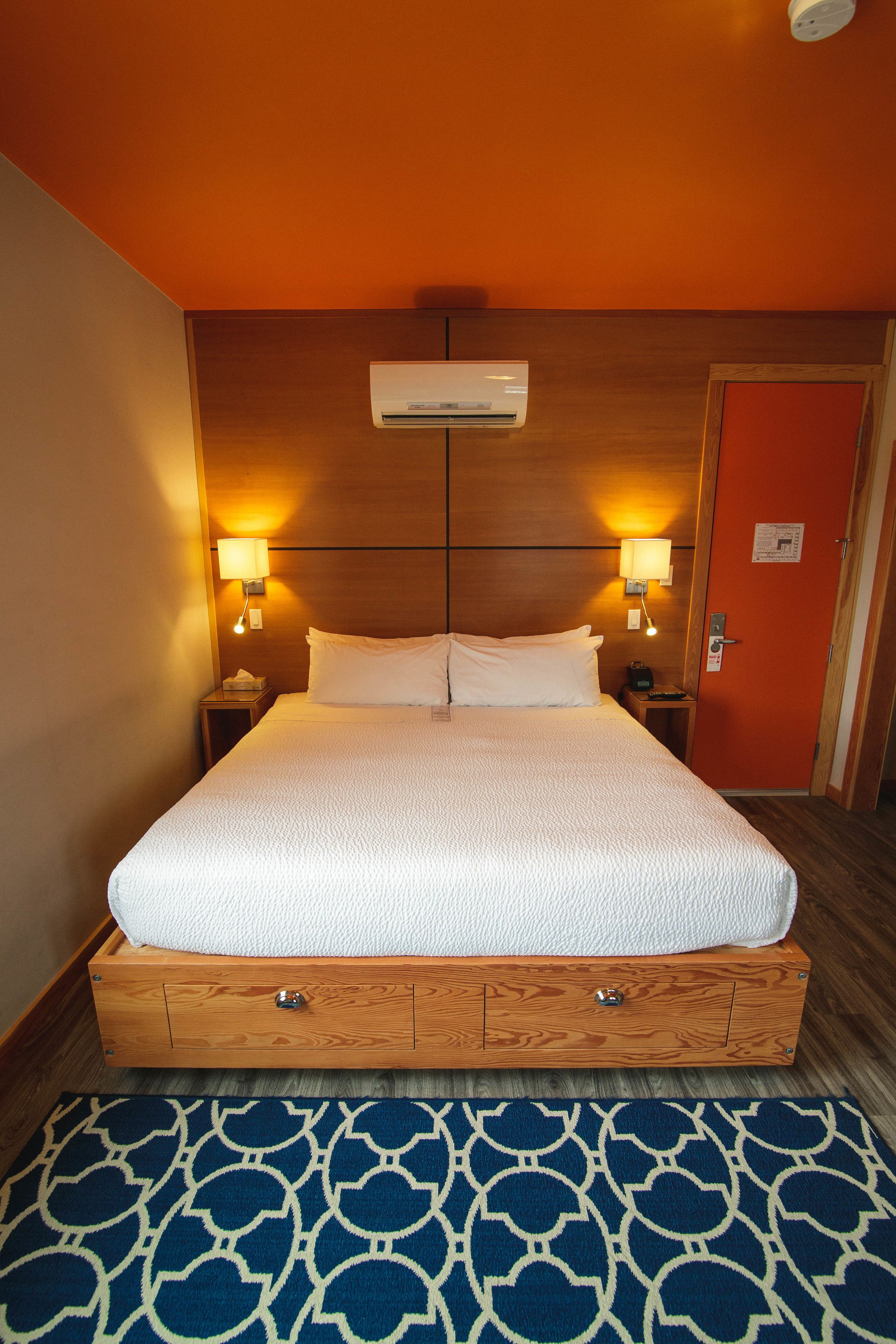 A hotel room with a large bed, wooden headboard, two bedside lamps, and an air conditioning unit above the bed.
