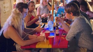 A group of people seated around a table with various items including bottles, cups, and a smartphone.