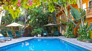 Una piscina con sillas de descanso y sombrillas, rodeada de vegetación exuberante y un edificio con balcones.