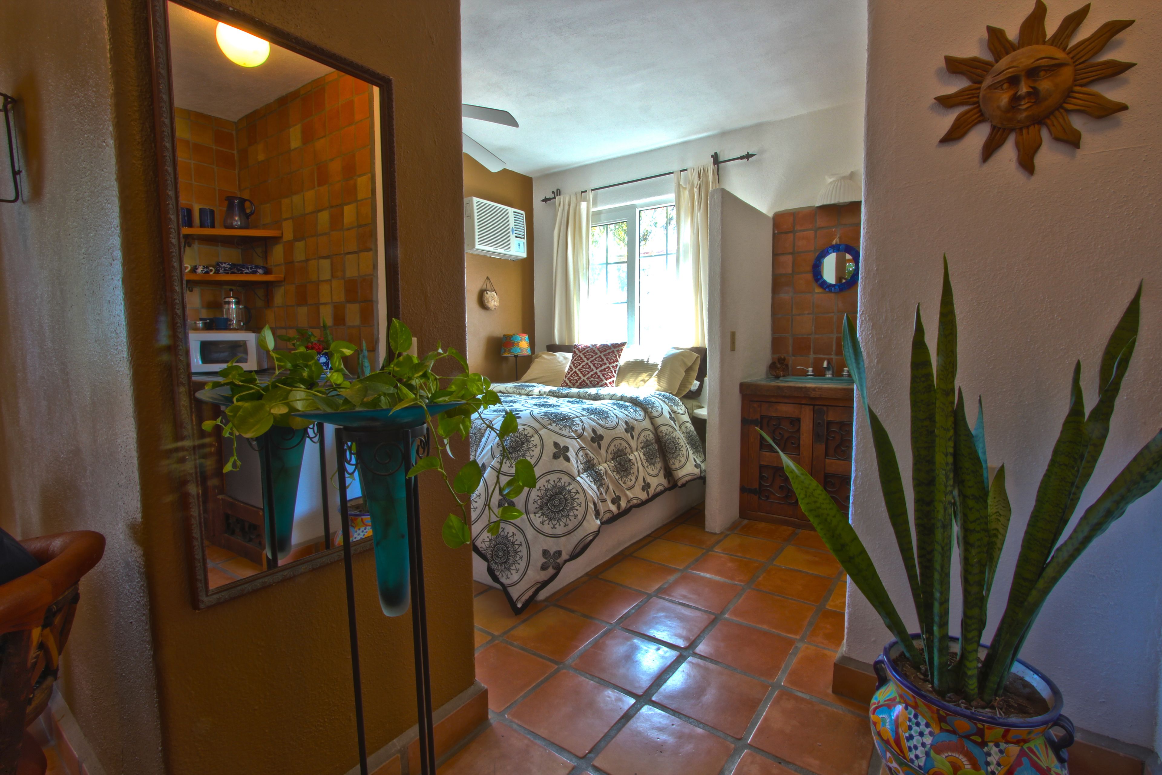 A bedroom with a bed, a window with curtains, a potted plant, and a bathroom visible through an open door.