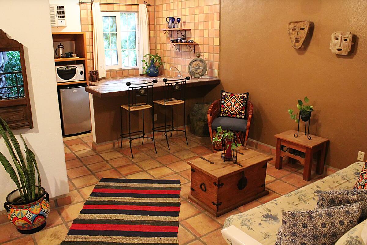 Eine Küche mit Mikrowelle, Kühlschrank und Barhocker. Ein Fenster mit Vorhang, ein Blumentopf und ein dekorativer Teller stehen auf der Arbeitsplatte.