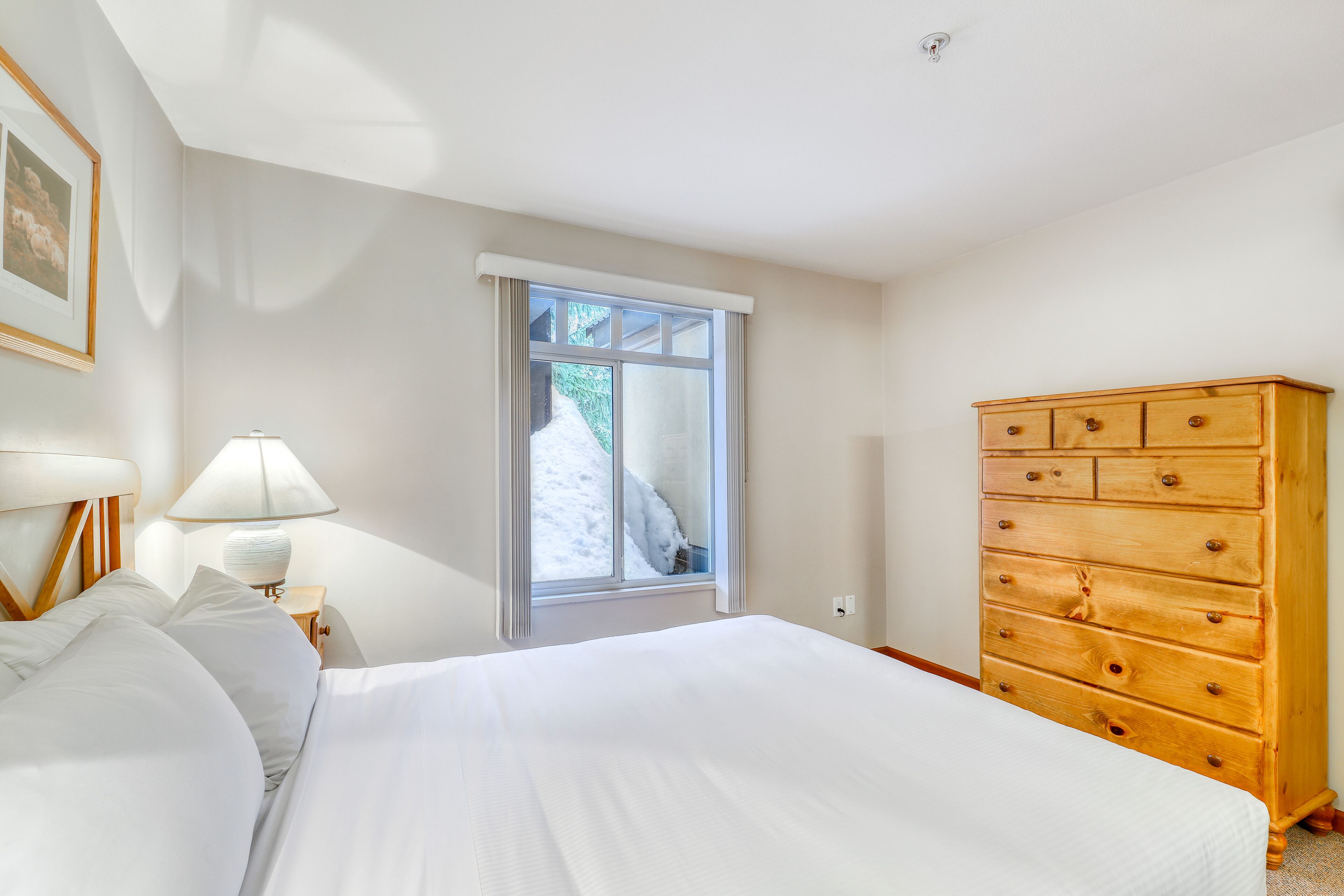Une chambre avec un grand lit, une commode en bois, une table de chevet avec une lampe et une fenêtre donnant sur un paysage enneigé.