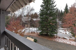 A balcony with a view of a snow-covered courtyard, a wooden bench, and a large evergreen tree.