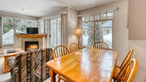 A cozy dining area with a wooden table and chairs, a fireplace, and large windows offering a view of the outdoors. - Stoney Creek Lagoon - Whistler Premier (Whistler)