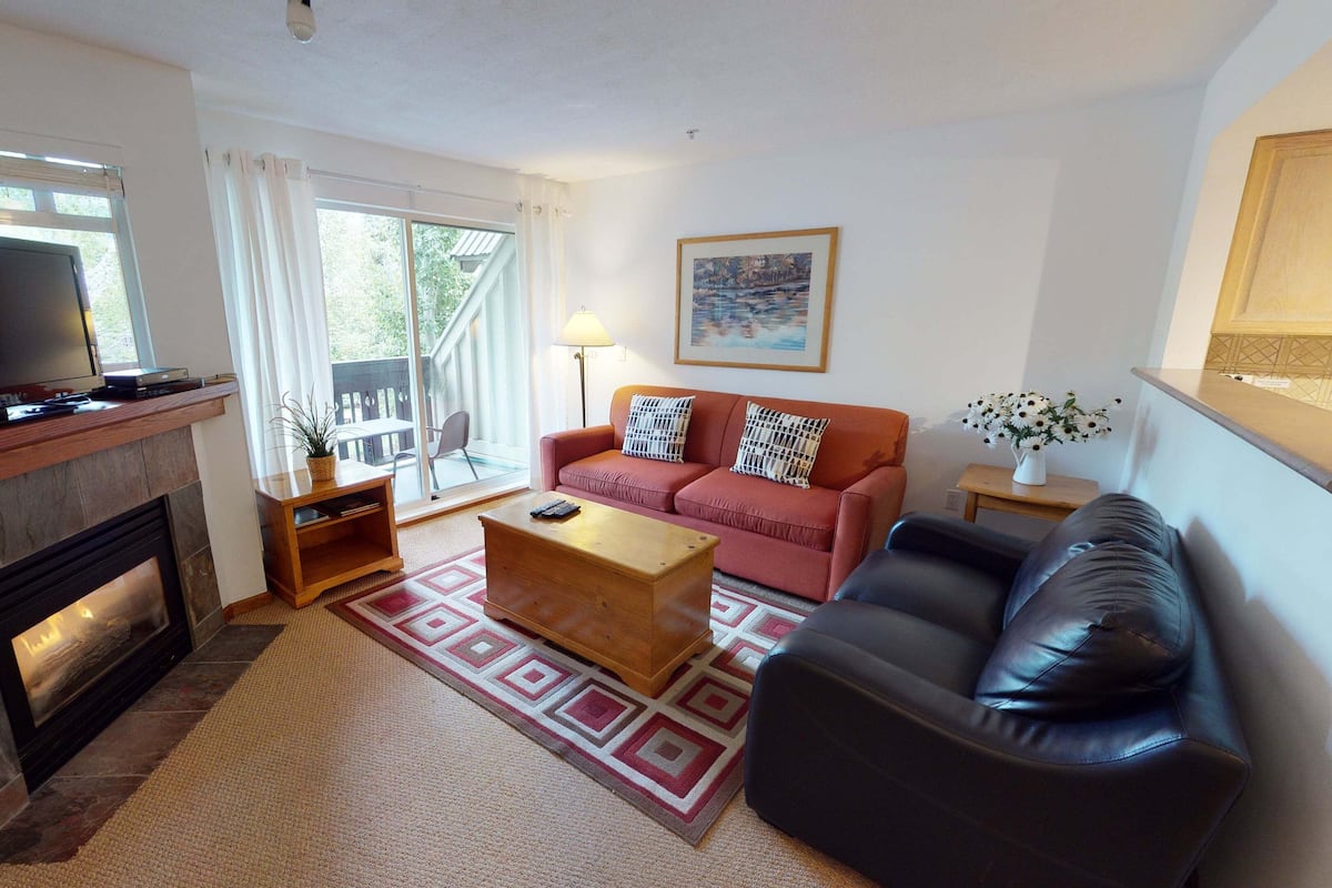 Ein Wohnzimmer mit Kamin, Fernseher, Sofa, Couchtisch und Blick ins Freie.