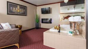 Habitación de hotel con un sofá de cuero marrón, un televisor de pantalla plana, una araña, una planta, un minibar y una bañera con una toalla.