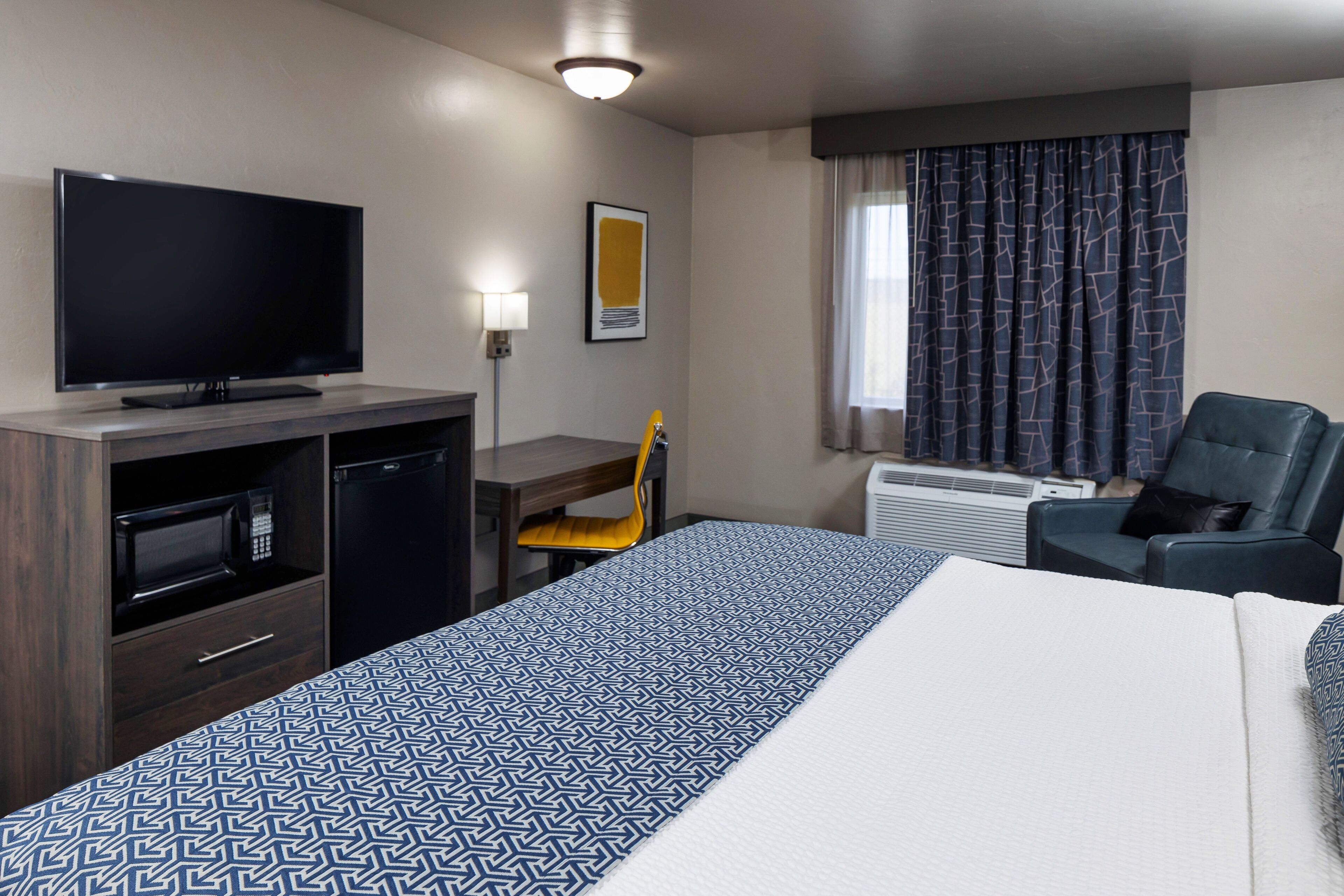 A hotel room with a bed, a desk with a chair, a television, a microwave, and an air conditioning unit.