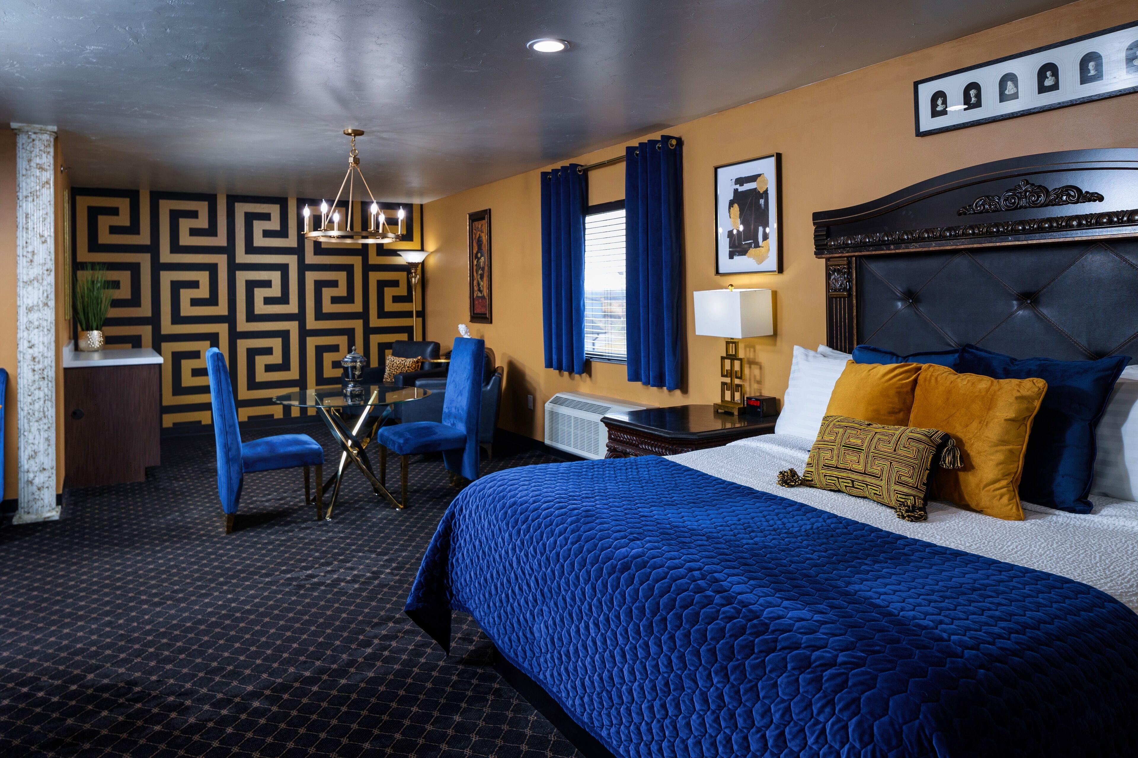A hotel room with a large bed, a desk, two chairs, a chandelier, and a patterned wall.