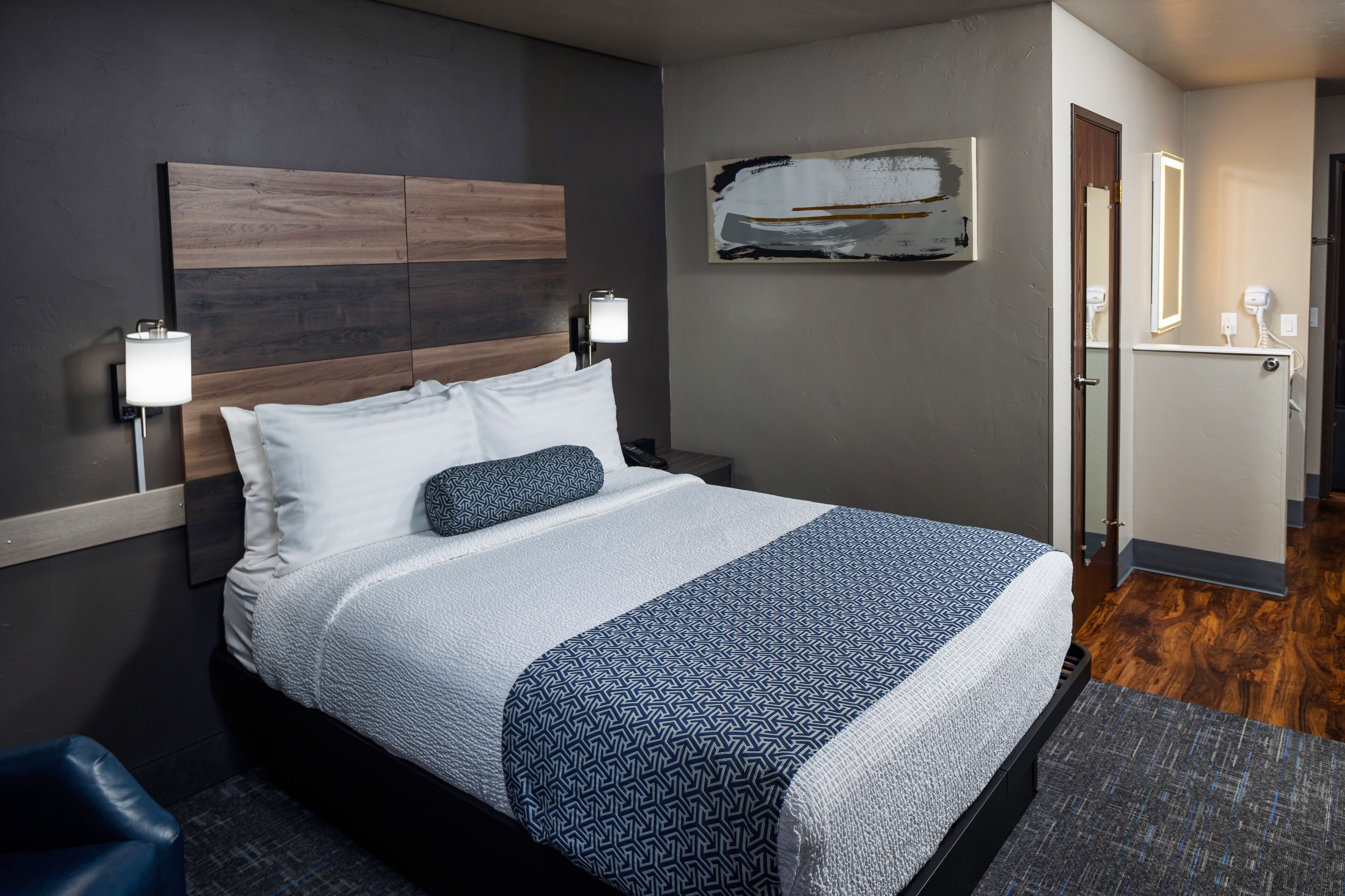 Habitación de hotel con una cama grande, una mesita de noche con lámpara, una obra de arte en la pared y una puerta que da al baño.