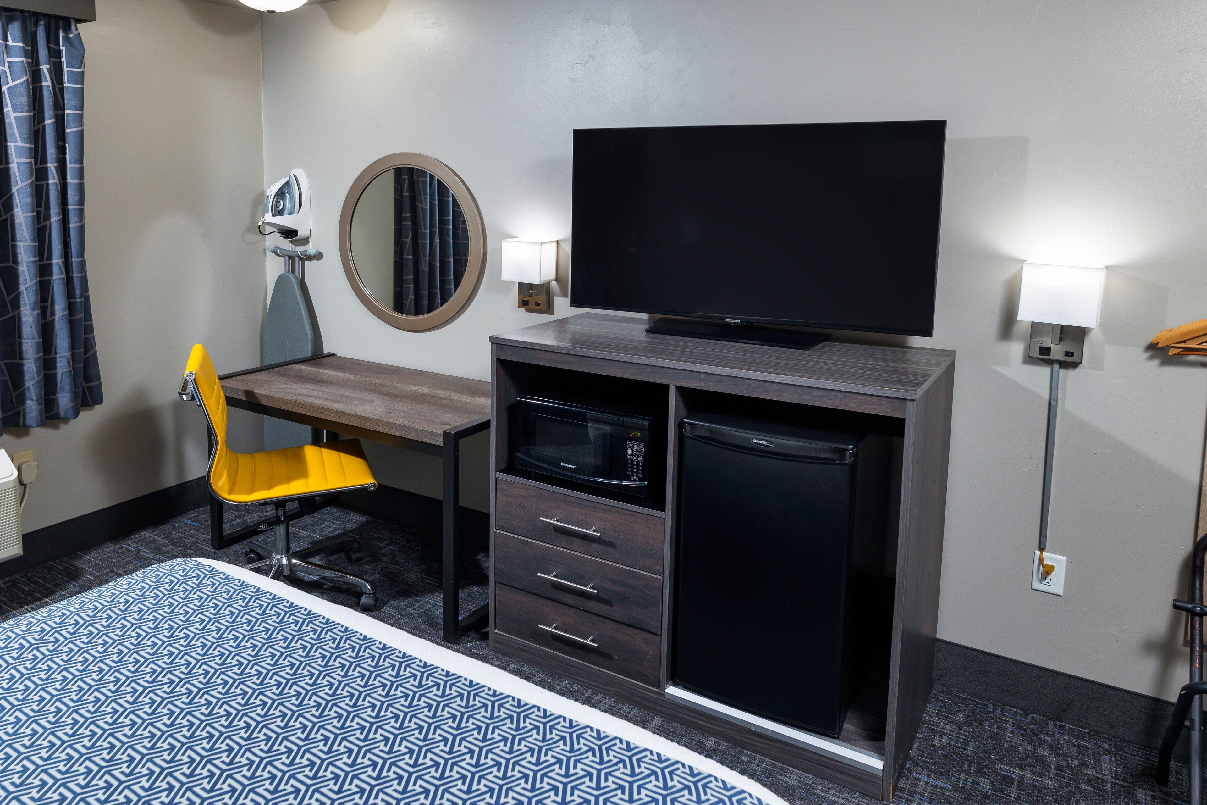 Quarto de hotel com cama, escrivaninha com cadeira, televisão, micro-ondas, frigorífico, espelho e duas luminárias de parede.