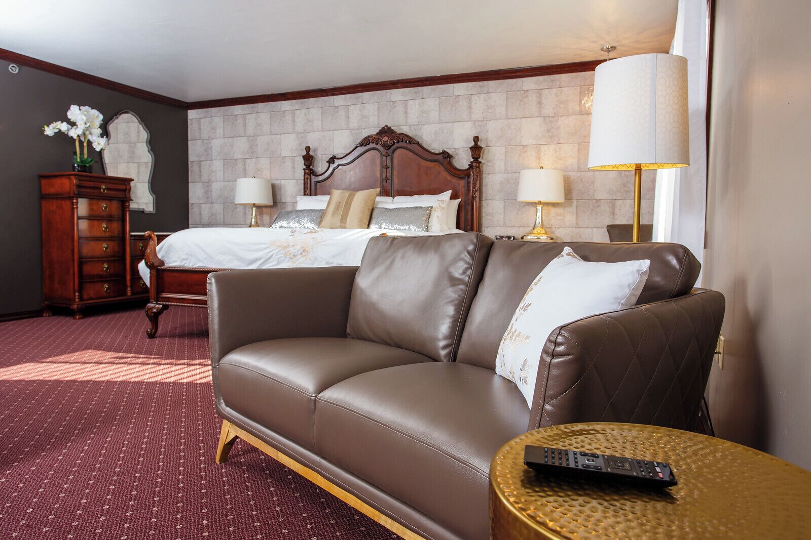 A hotel room with a brown leather sofa, a wooden dresser, a bed with white linens, two bedside lamps, and a round table with a remote control.