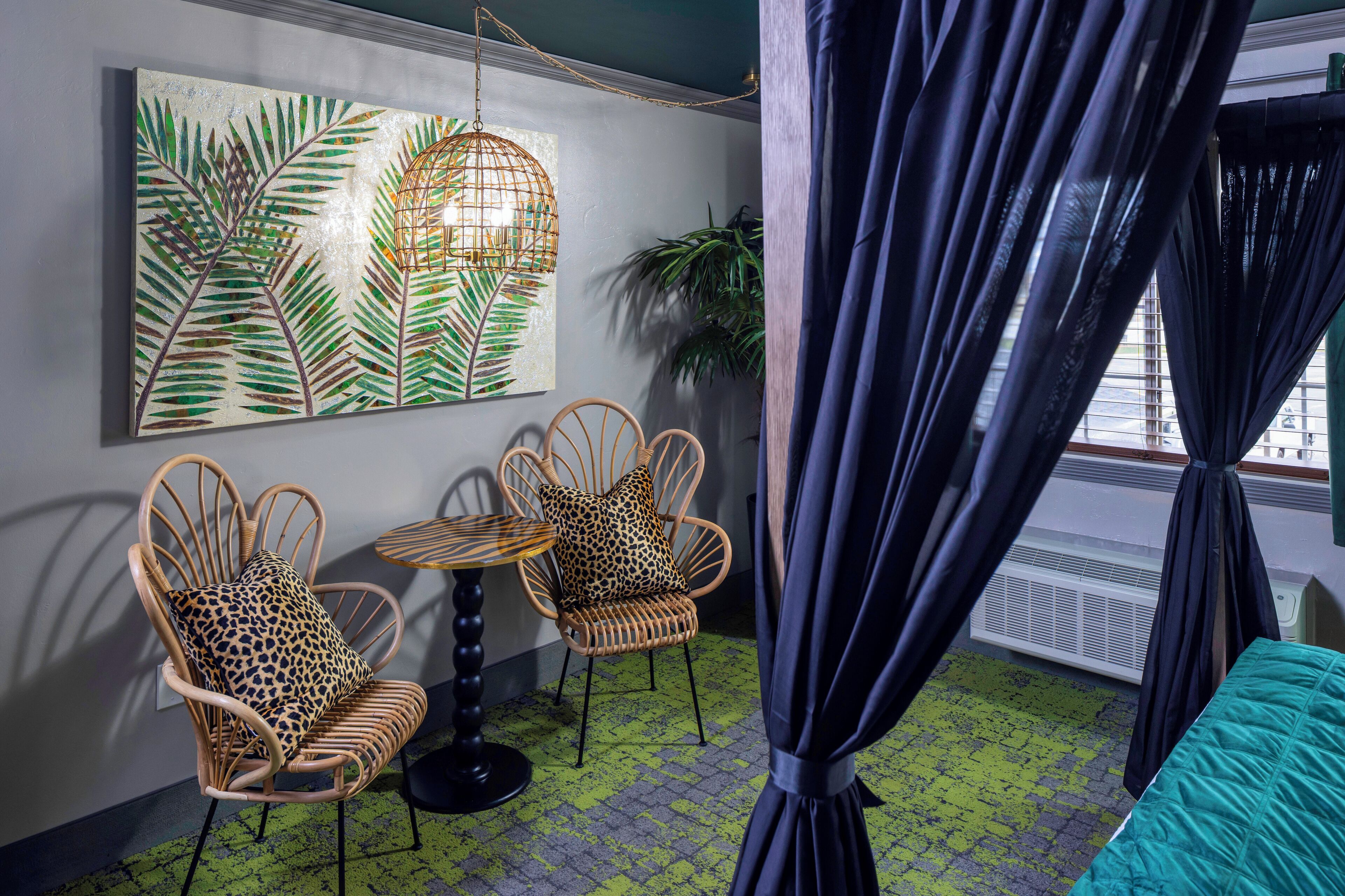 Een kamer met twee rieten stoelen, waarvan één met een luipaardprint kussen, een ronde tafel, een vloer met een groen patroon en een groot schilderij met bladeren aan de muur.