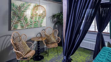 Een kamer met twee rieten stoelen, waarvan één met een luipaardprint kussen, een ronde tafel, een vloer met een groen patroon en een groot schilderij met bladeren aan de muur.