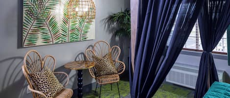 A room with two wicker chairs, one with a leopard print cushion, a round table, a green patterned floor, and a large leaf-patterned painting on the wall.