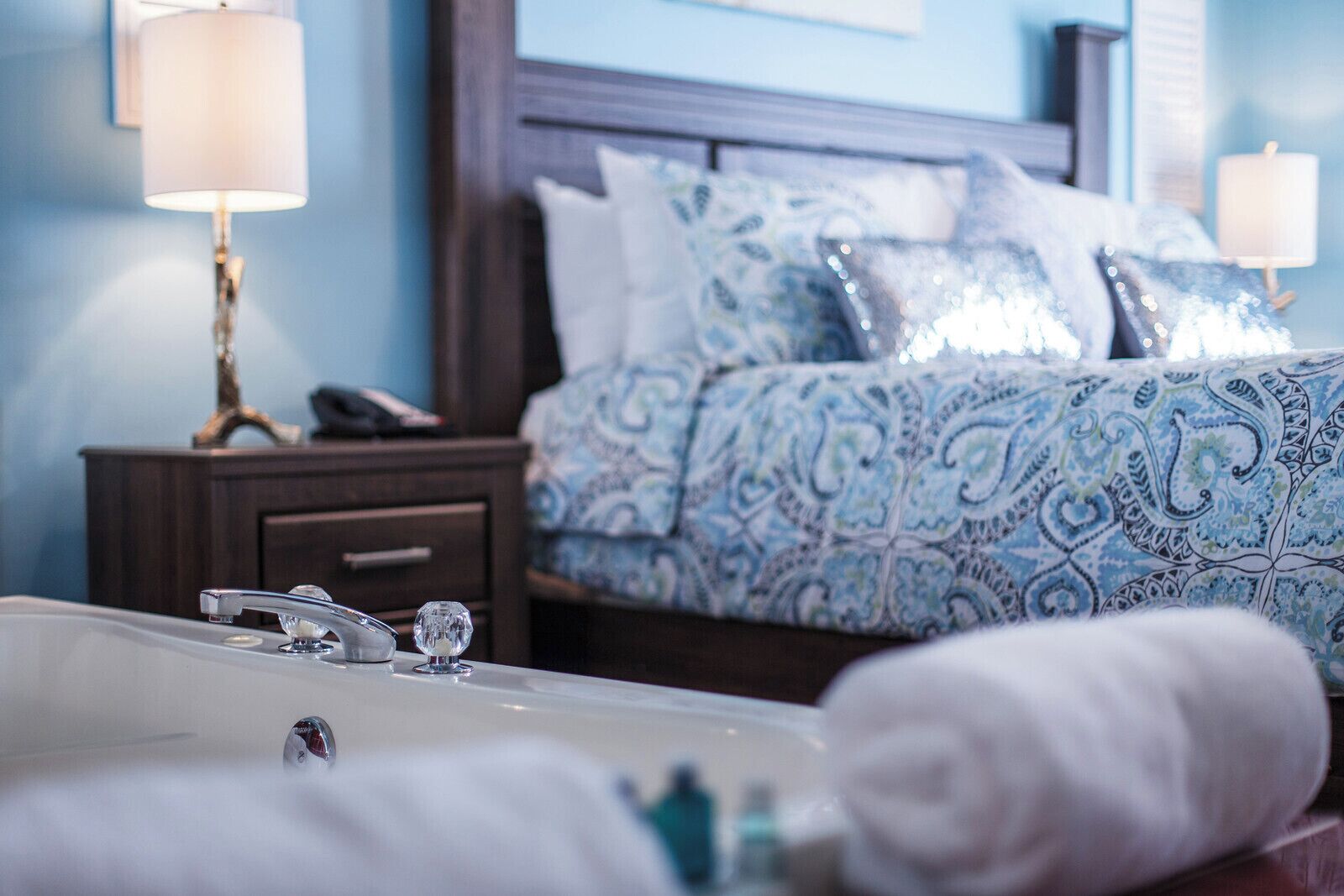 A hotel room with a large bed, a nightstand, a bathroom with a bathtub, and a wall-mounted lamp.