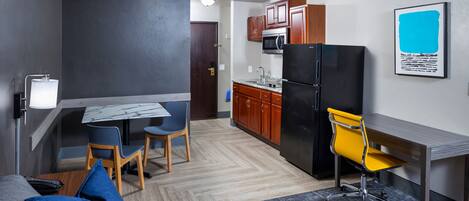 A compact kitchen with a microwave, refrigerator, and wooden cabinets. There is a dining area with a table and chairs, and a sofa with a cushion.