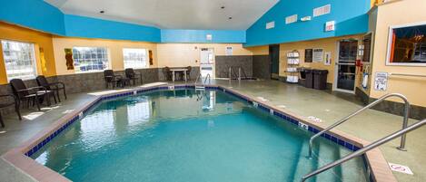 Una piscina interior con agua azul cristalina, rodeada por un borde de azulejos y una barandilla de seguridad. Cerca de la piscina hay sillas y una mesa, y una puerta que da a otra habitación.