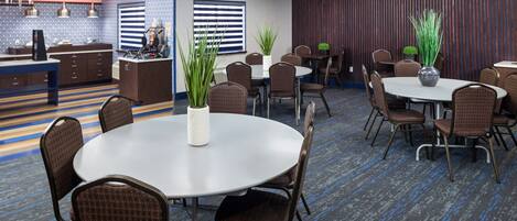 Un salón de conferencias con mesas y sillas redondas, plantas en macetas y una zona de cocina al fondo.