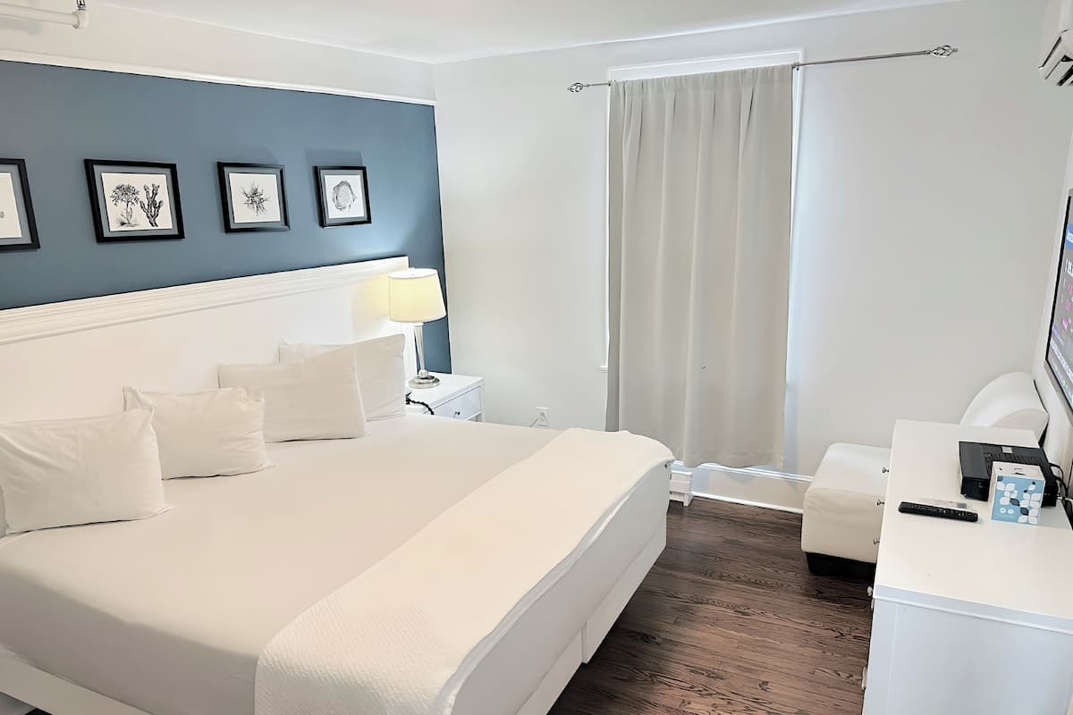 Hotelkamer met een groot bed, een nachtkastje met lamp, een bureau met computer en afstandsbediening, een stoel en een raam met gordijnen.