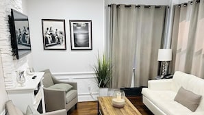 A modern living room with a white sofa, two armchairs, a wooden coffee table, and a white fireplace. There are framed pictures on the wall and a potted plant.