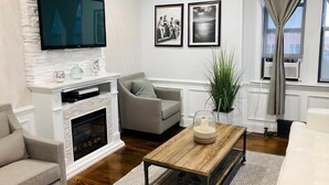 A living room with a fireplace, a flat-screen TV mounted on a white brick wall, a grey sofa, a wooden coffee table, and a large potted plant.
