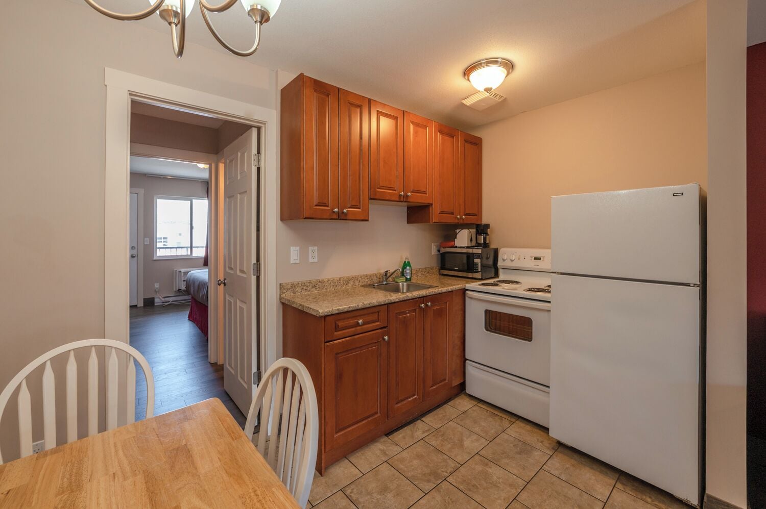 A kitchen with wooden cabinets, a white refrigerator, a microwave, and a stove. There is a dining table with chairs and a window in the background.