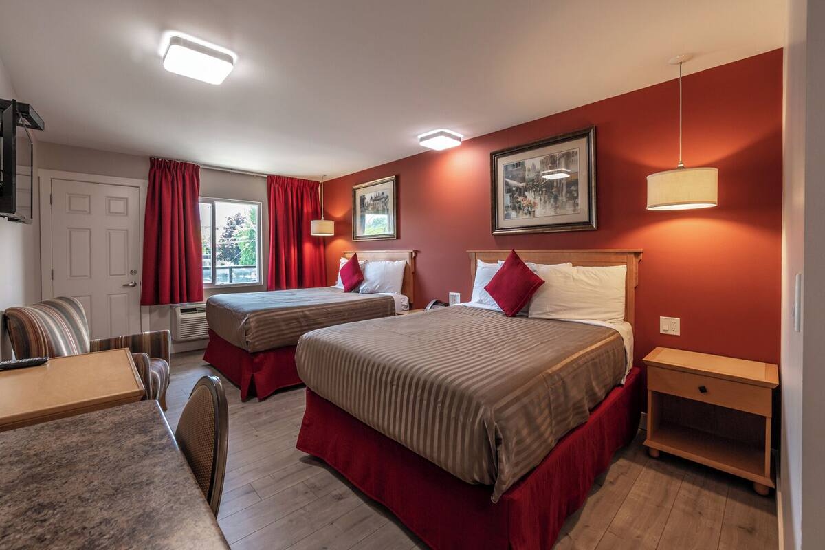 Habitación de hotel con dos camas, un escritorio y una silla. Hay dos ventanas con cortinas rojas, un cuadro en la pared y una lámpara sobre la mesita de noche.