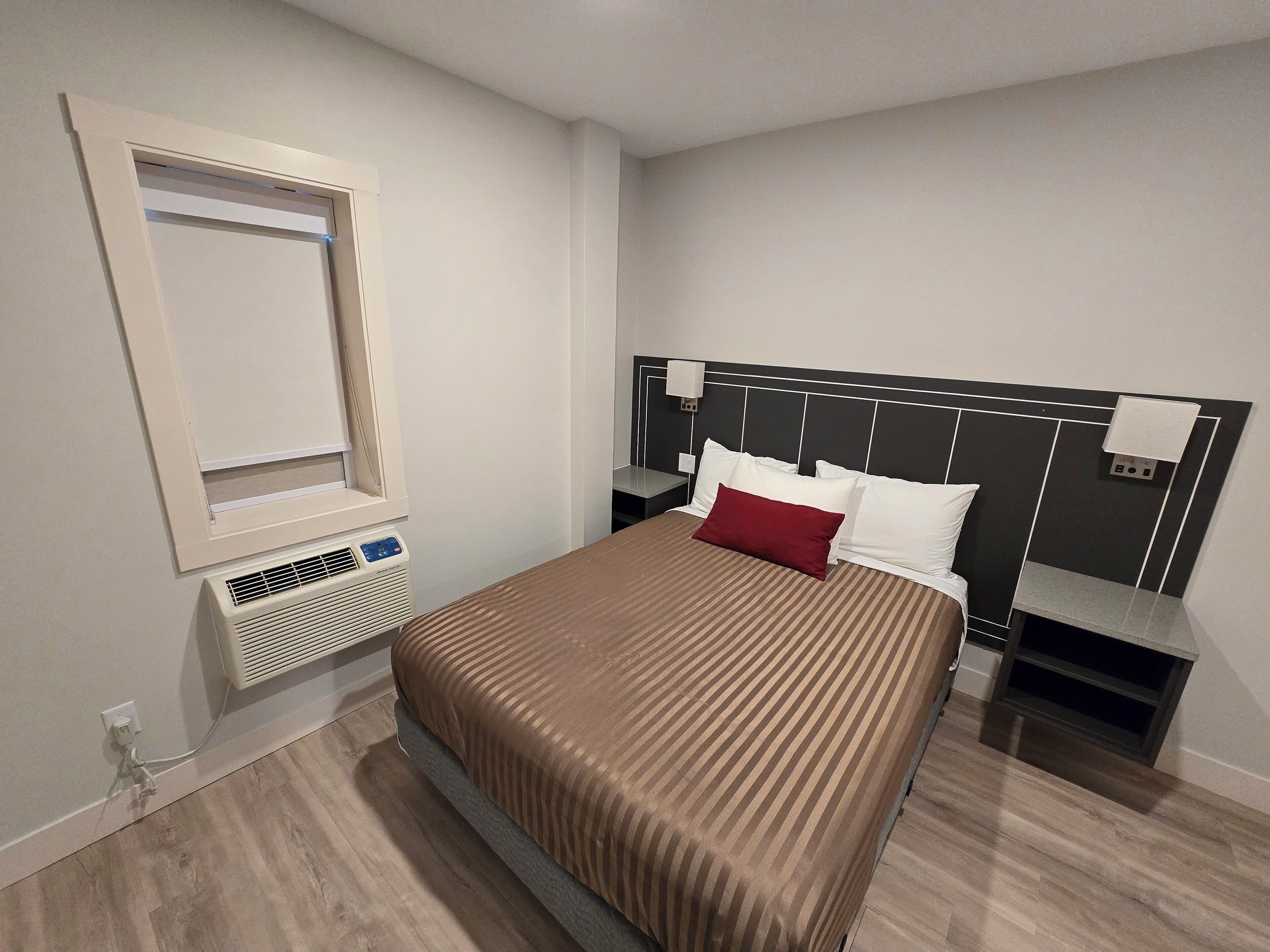 A hotel room with a bed, a nightstand, a bedside table, a wall-mounted air conditioner, and a window with blinds.