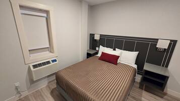 A hotel room with a bed, a nightstand, a bedside table, a wall-mounted air conditioner, and a window with blinds.