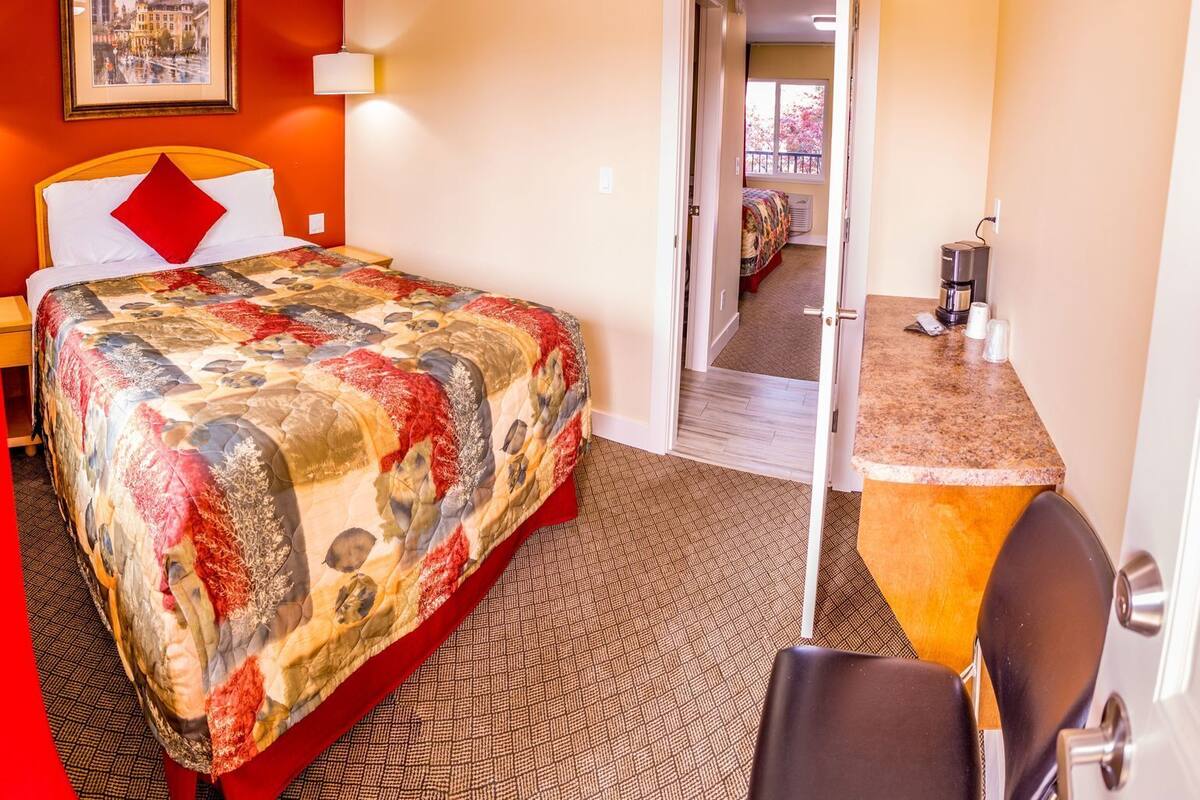 Habitación de hotel con cama, escritorio, silla, cafetera y puerta que da a otra habitación.
