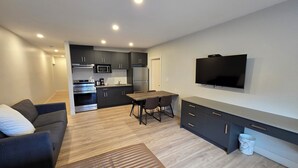 Un apartamento moderno con cocina, comedor y sala de estar. Cuenta con un sofá, una mesa de comedor con sillas, un televisor montado en la pared y un cubo de basura.