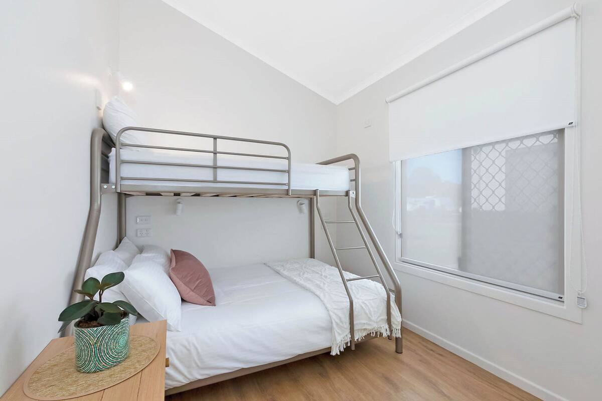 A bunk bed with a white mattress and a single pink pillow, a small green plant on a wooden side table, and a window with a white blind.