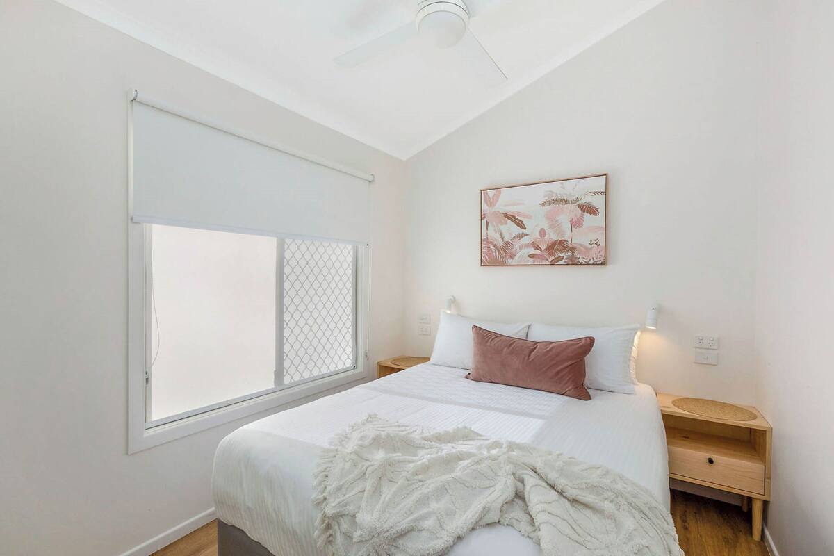 A bedroom with a bed, a window with a white blind, a ceiling fan, a bedside table with a lamp, and a wall-mounted artwork.