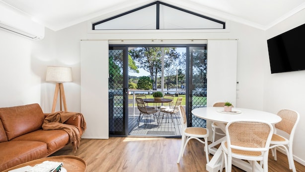 A modern living room with a brown leather sofa, a round white table with wicker chairs, and a large sliding glass door leading to an outdoor patio with a table and chairs.