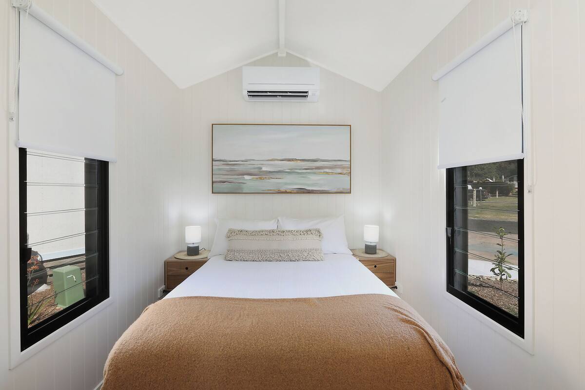 A bedroom with a bed, two bedside tables with lamps, a painting above the bed, and two windows with blinds.