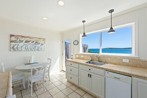 A kitchen with a view of the sea, a dining area with chairs, a clock, and a painting on the wall.