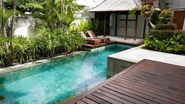 Piscina con cubierta de madera y sillones de descanso, rodeada de exuberante follaje.