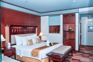 Habitación de hotel con una cama grande, mesitas de noche, un banco, una lámpara, cabecera de madera, repisa en la pared y una puerta que da a otra habitación.