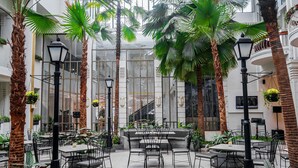 A spacious indoor courtyard with multiple dining tables and chairs, surrounded by tall palm trees and potted plants. - Hotel Savoy Homann (Bandung)