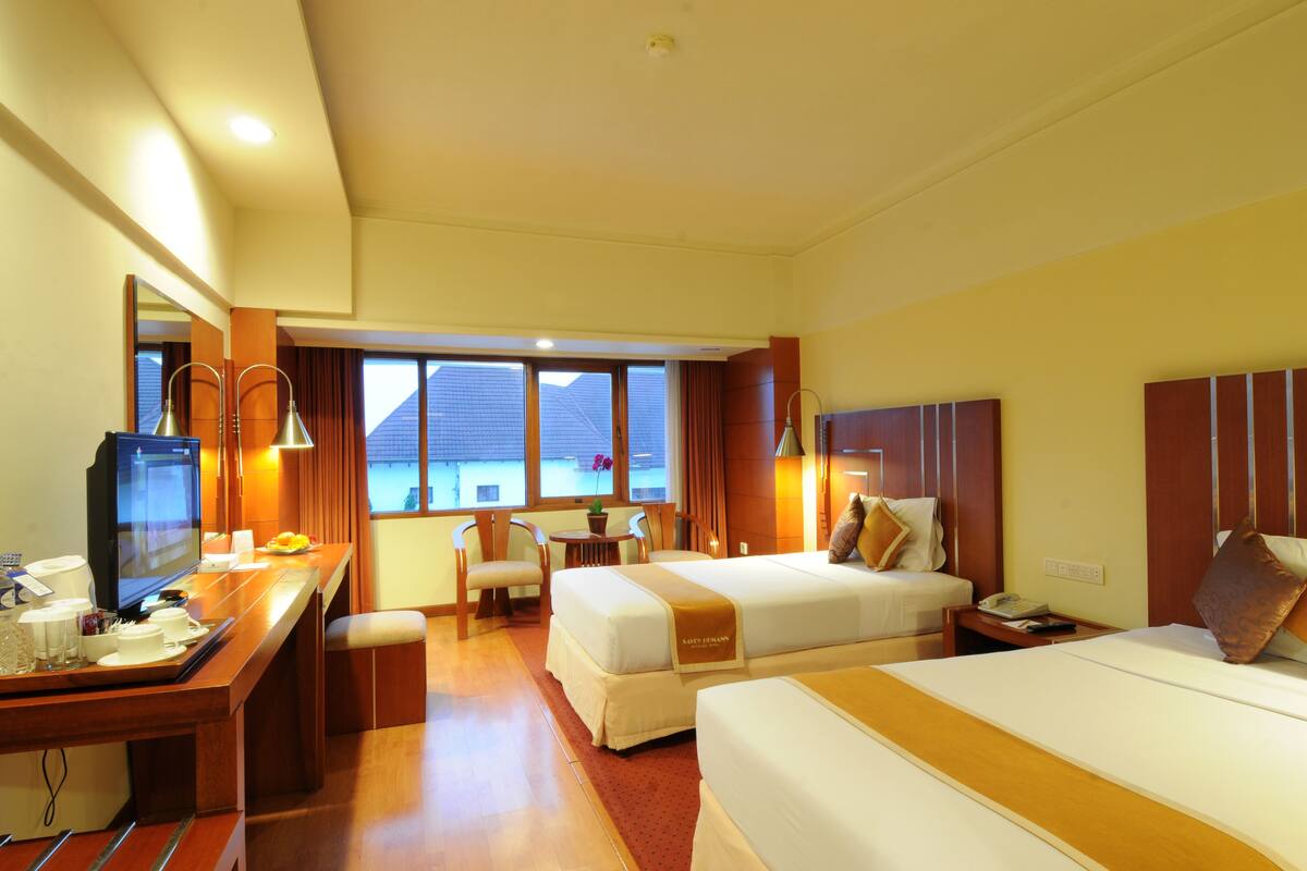 A hotel room with two beds, a desk with a TV, a small table with chairs, and a view of buildings through large windows.
