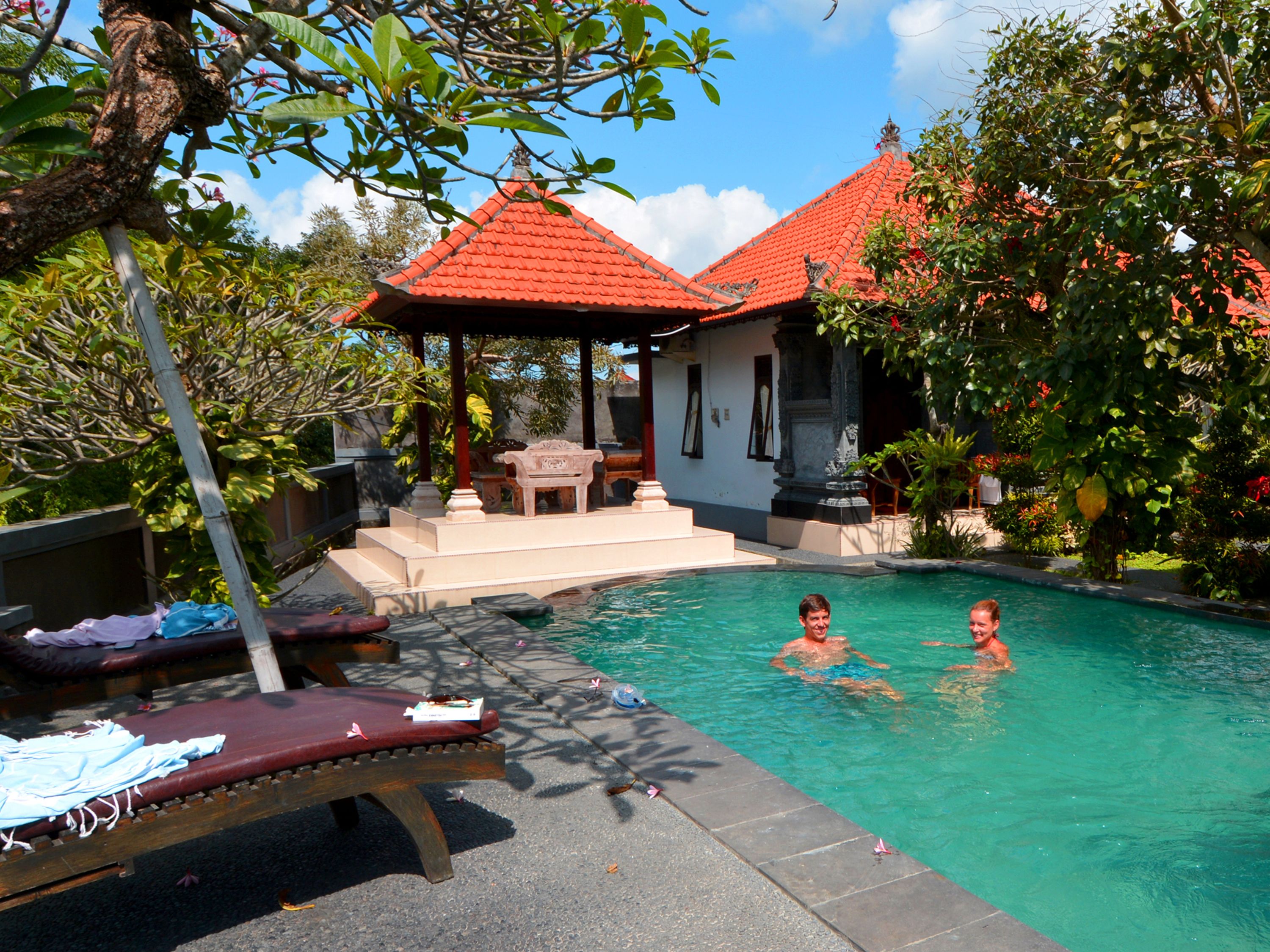 A swimming pool with two people in the water, surrounded by lush greenery and a building with a red roof.