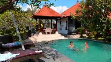 A swimming pool with two people in the water, surrounded by lush greenery and a building with a red roof.