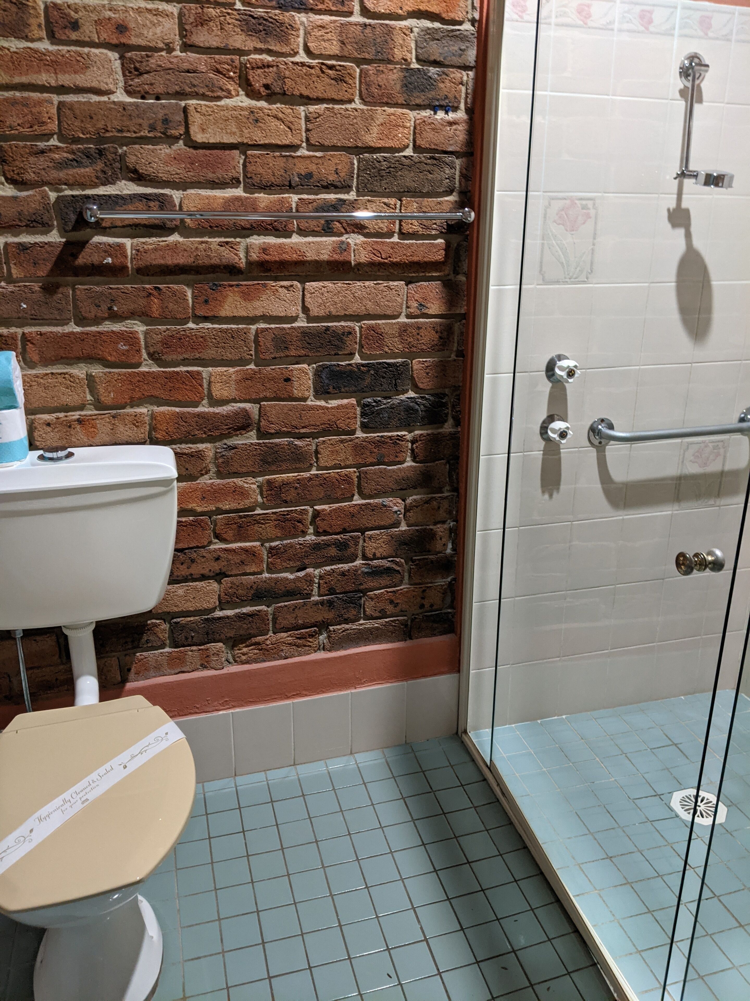 A bathroom with a brick wall, a toilet, a shower with a glass door, and tiled flooring.