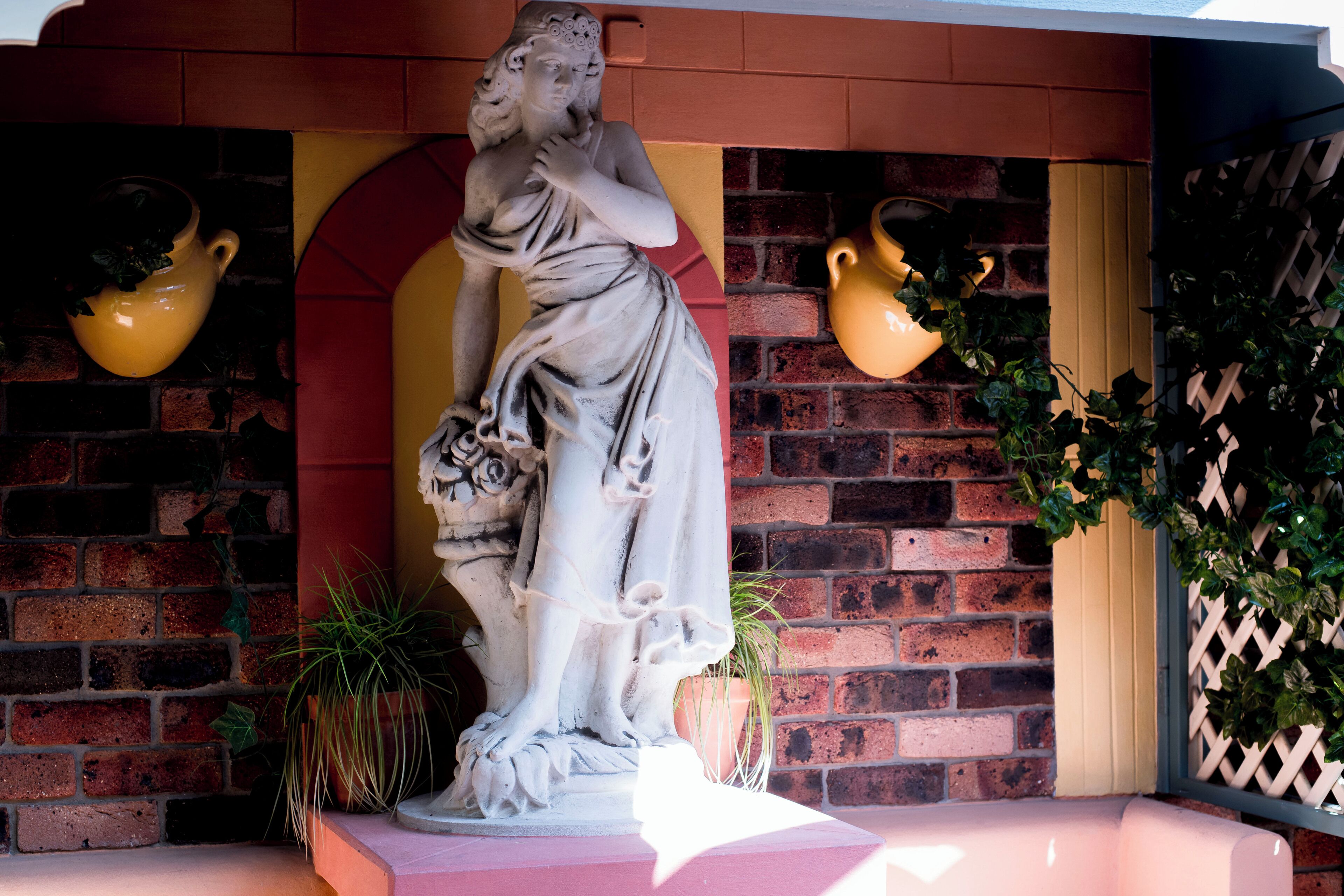 Una estatua de una mujer sosteniendo un libro, frente a un muro de ladrillo con macetas amarillas y una planta en maceta cerca.